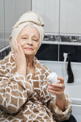 A seventy year old woman with a towel on her head, in a bathrobe sits on a chair in her bathroom...