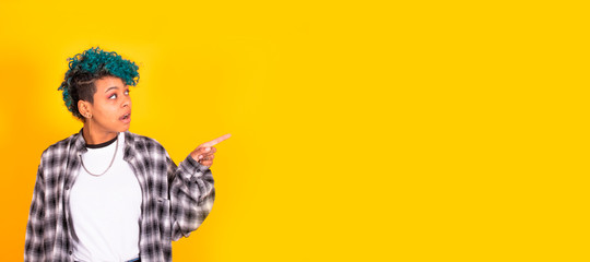 young african american woman with blue curly hair pointing at yellow background