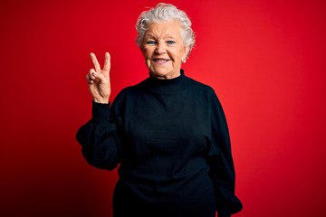 Senior beautiful woman wearing casual sweater standing over isolated red background smiling with happy face winking at the camera doing victory sign. Number two.