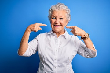 Senior beautiful woman wearing elegant shirt standing over isolated blue background smiling cheerful showing and pointing with fingers teeth and mouth. Dental health concept.