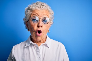 Senior beautiful woman wearing elegant shirt and glasses over isolated blue background afraid and shocked with surprise expression, fear and excited face. - obrazy, fototapety, plakaty