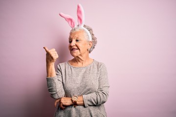 Senior beautiful woman wearing bunny ears standing over isolated pink background smiling with happy face looking and pointing to the side with thumb up.