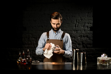 Professional bartender standing on the bar counter rubs the glass cup with a rag