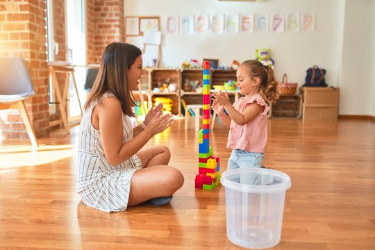 Beautiful teacher and blond toddler girl building tower using plastic blocks at kindergarten