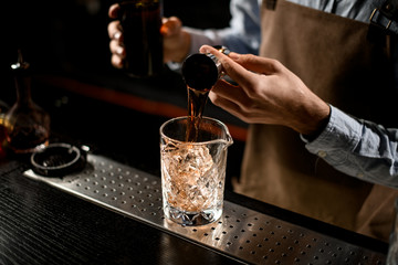 Professional bartender pouring a golden brown alcoholic drink from the steel jigger to a glass