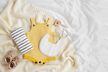 Yellow bodysuit, bib, baby boots and toy on knitted blanket. Set of  kids clothes and accessories  on bed. Fashion newborn. Flat lay, top view