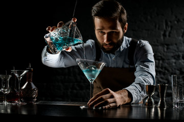 Male bartender pouring a blue color alcoholic cocktail from the measuring cup with strainer to a martini glass
