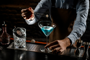 Professional bartender decorating a blue alcoholic cocktail in a martini glass with a spikelet by tweezers