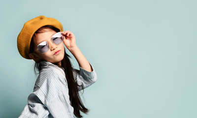 Little asian kid in sunglasses, oversized shirt dress, brown beret, boots. She kissing you, posing on blue background. Full length