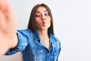 Beautiful woman wearing denim shirt make selfie by camera over isolated white background making fish face with lips, crazy and comical gesture. Funny expression.
