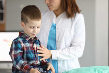 Portrait of child sitting in modern clinic office and looking down in gloomy mood. Female...