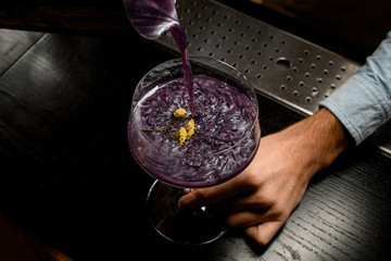 Bartender pouring purple alcoholic drink to the big cocktail glass with a yellow flower
