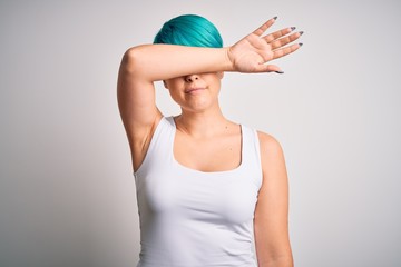 Young beautiful woman with blue fashion hair wearing casual t-shirt over white background covering eyes with arm, looking serious and sad. Sightless, hiding and rejection concept