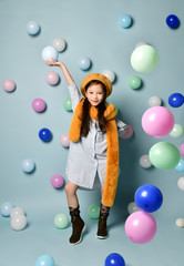 Fototapeta premium Little asian kid in oversized shirt dress, brown beret and scarf, boots. She smiling, posing with balloons on blue background.