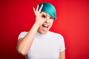 Young beautiful woman with blue fashion hair wearing casual t-shirt over red background doing ok gesture with hand smiling, eye looking through fingers with happy face.