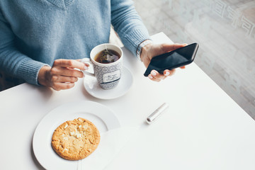 Woman is having a breakfast in a modern cafe, drinks rooibos and eats oat cookie. Uses cellular phone, checks updates, reads article, creates new publication on own web page. 