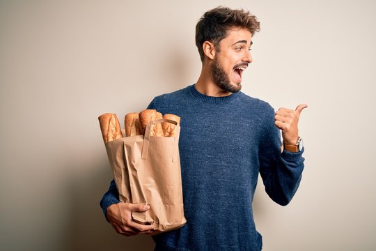 Young man holding paper bag with bread standing over isolated red bakground pointing and showing with thumb up to the side with happy face smiling