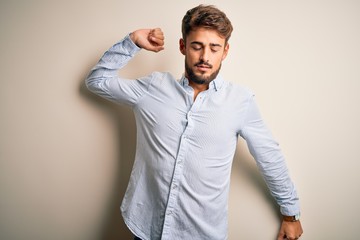 Young handsome man with beard wearing striped shirt standing over white background stretching back, tired and relaxed, sleepy and yawning for early morning