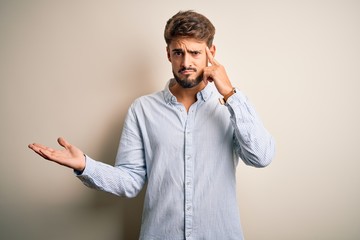 Young handsome man with beard wearing striped shirt standing over white background confused and...