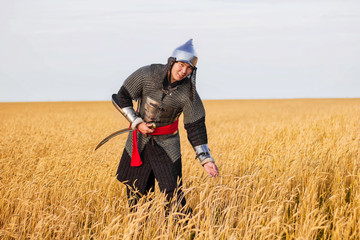 A nomad warrior in oriental armor with a saber and a helmet from the 16th and 18th centuries stands knee-deep in a wheat field.