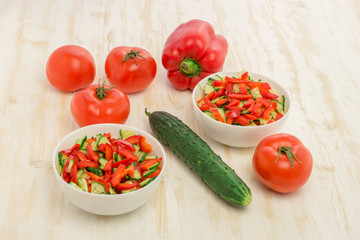 Vitamin salad of ripe, red tomatoes and fresh, green cucumbers in glass white bowl on light wooden table. Ingredients for salad. Vegetables. Concept of healthy eating