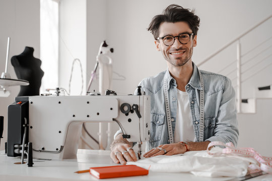 Creative creative work, attractive seamstress man sitting at the sewing machine and smiling. Sewing, design work, tailoring studio, tailor, designer clothes, manufactory, in process of creative work