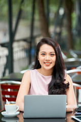 Vertical portrait of beautiful Asian woman wearing pale pink dress sitting at cafe table outdoors looking at camera smiling