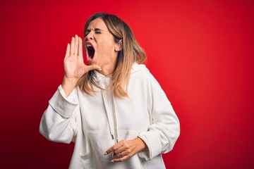 Middle age beautiful sportswoman wearing casual sweatshirt over isolated red background shouting and screaming loud to side with hand on mouth. Communication concept.