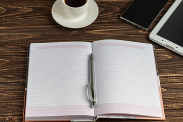 A wooden office desk with many things on it. Diary with pen, Samsung phone. White cup of coffee....