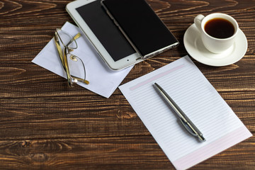 A wooden office desk with many things on it. Diary with pen, Samsung phone. White cup of coffee. Top view with copy space,