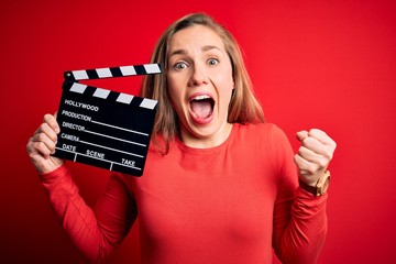 Young beautiful blonde clapper woman holding clapboard over isolated red background screaming proud...
