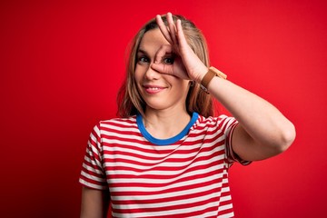 Young beautiful blonde woman wearing casual striped t-shirt over isolated red background doing ok gesture with hand smiling, eye looking through fingers with happy face.