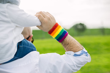 Adult man wearting a gay pride rainbow flag wristband and carrying his adopted son in the park. LGTB and equality concept