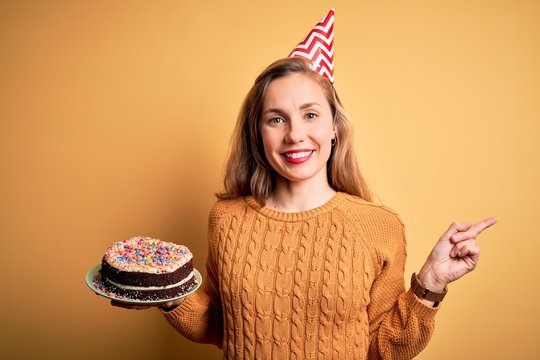 Young beautiful blonde woman holding birthday cake over isolated yellow background very happy pointing with hand and finger to the side