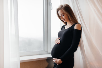 A beautiful pregnant girl is standing in a room near the window