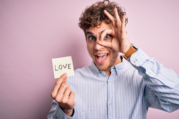 Young blond romantic man with curly hair holdiing paper with love message for valentines day with happy face smiling doing ok sign with hand on eye looking through fingers