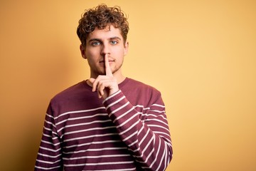 Young blond handsome man with curly hair wearing casual striped sweater asking to be quiet with finger on lips. Silence and secret concept.
