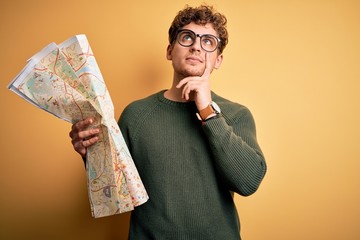 Young blond tourist man on vacation with curly hair holding city map over yellow background serious...