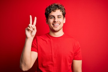 Naklejka premium Young blond handsome man with curly hair wearing casual t-shirt over red background showing and pointing up with fingers number two while smiling confident and happy.