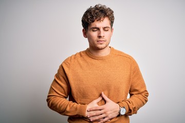Young blond handsome man with curly hair wearing casual sweater over white background with hand on stomach because indigestion, painful illness feeling unwell. Ache concept.