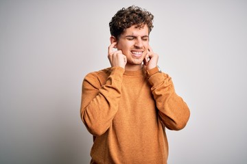 Young blond handsome man with curly hair wearing casual sweater over white background covering ears with fingers with annoyed expression for the noise of loud music. Deaf concept.