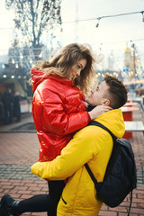 young romantic couple in love hugging, wearning in bright yellow and red down jackets. Handsome man carrying young attractive woman while walking on the street.