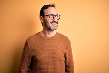 Middle age hoary man wearing brown sweater and glasses over isolated yellow background looking away to side with smile on face, natural expression. Laughing confident.