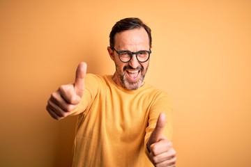 Fototapeta premium Middle age hoary man wearing casual sweater and glasses over isolated yellow background approving doing positive gesture with hand, thumbs up smiling and happy for success. Winner gesture.