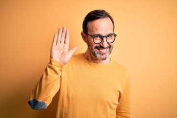 Middle age hoary man wearing casual sweater and glasses over isolated yellow background Waiving saying hello happy and smiling, friendly welcome gesture