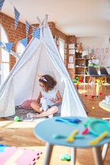 Beautiful toddler wearing glasses and unicorn diadem sitting on the floor inside tipi holding doll and smiling at kindergarten