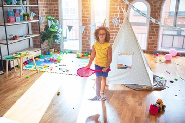 Beautiful toddler standing wearing glasses and unicorn diadem playing with tennis racket at kindergarten