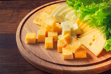 Square pieces of cheese with herbs on a round wooden board.