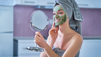 Smiling young happy healthy female in bath towel with green cleansing face clay mask and small...