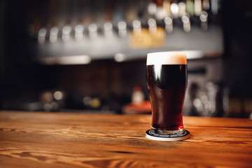 Glass of dark beer craft with foam on wooden bar counter, pub blurred background
